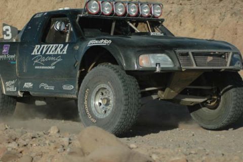 1990 Ford Bronco - black off-road race truck number 3 with Riviera Racing graphics and a six-light bar races on a rocky course.