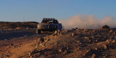 1990 Ford Bronco - silver off-road race truck topped with a light bar crests a rocky rise, trailing dust in low evening light.