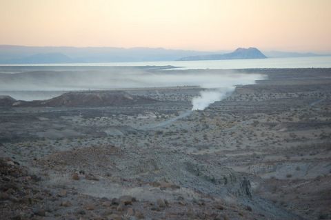 1990 Ford Bronco - faint dust trail rises from a vehicle crossing a hazy desert valley with water and mountains in the distance at dawn.