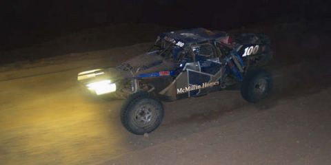 1990 Ford Bronco - off-road buggy number 100 marked McMillin Homes races at night with its headlights blazing.