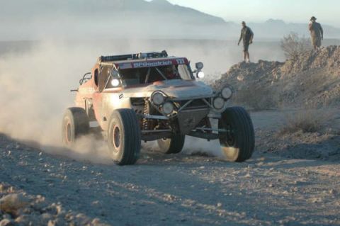 1990 Ford Bronco - orange open-cockpit tube-frame off-road race buggy speeds along a dusty desert track as people watch.