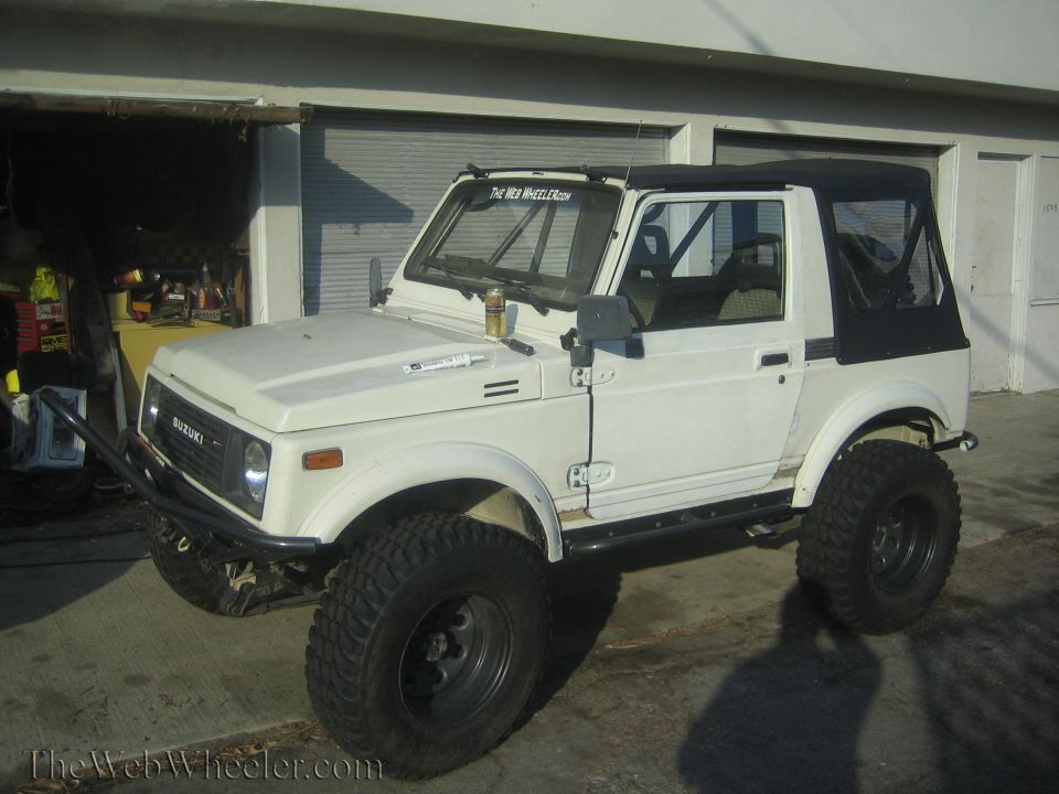 White 1987 Suzuki Samurai with aftermarket bumper and side steps parked in front of a garage