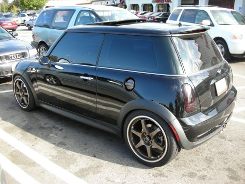 1998 Ford Crown Victoria - black Mini Cooper hardtop with bronze alloy wheels parked in a lot, seen from the rear three-quarter angle.