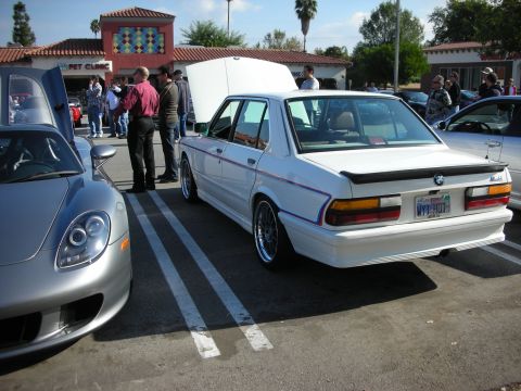 1998 Ford Crown Victoria - white BMW E28 sedan with a rear spoiler and Florida plates sits next to a silver Porsche Carrera GT at a strip-mall car meet.