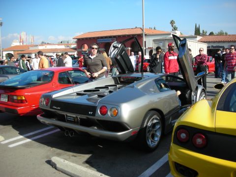 1998 Ford Crown Victoria - silver Lamborghini Diablo VT with its scissor doors raised draws onlookers at a parking-lot car meet, flanked by a red and yellow sports ca…