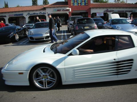 1998 Ford Crown Victoria - white Ferrari Testarossa with prominent side strakes rolls through a strip-mall parking lot lined with other show cars.