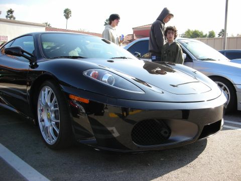 1998 Ford Crown Victoria - black Ferrari F430 coupe with five-spoke wheels and a prancing-horse fender badge is parked at a car meet as people walk by.