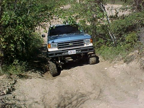 1990 Ford Bronco - A blue pickup truck navigates a rough, wooded trail.