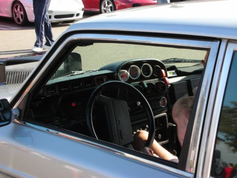 1998 Ford Crown Victoria - Through the open window of an older sedan, an aftermarket gauge cluster and three-spoke steering wheel are visible with a driver inside.