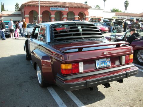 1998 Ford Crown Victoria - maroon Volvo 240 coupe with a rear-window louver and flared fenders is parked rear-out at a busy strip-mall car meet.
