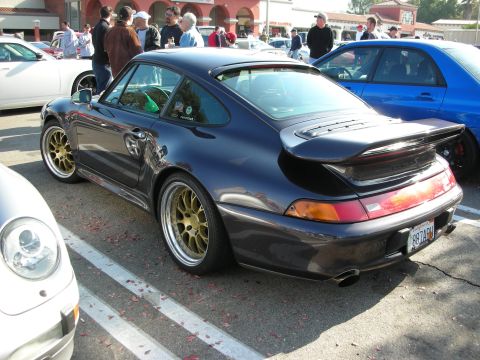1998 Ford Crown Victoria - dark Porsche 993 with a whale-tail spoiler and gold wheels is parked among other cars at an outdoor car meet.