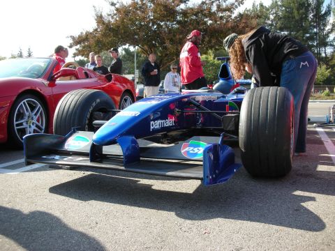 1998 Ford Crown Victoria - blue single-seat open-wheel formula race car carrying Parmalat sponsorship sits in a parking lot next to a red Ferrari.