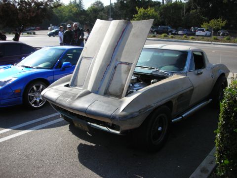 1998 Ford Crown Victoria - unpainted mid-1960s Corvette coupe sits with its tilt-up front clip raised, beside a blue convertible, at an outdoor car meet.