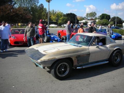 1998 Ford Crown Victoria - unpainted mid-1960s Corvette coupe rolls through a crowded car meet past red Ferraris and a blue open-wheel race car.