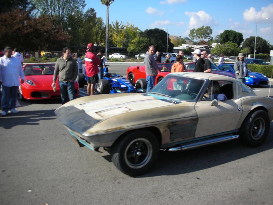 Bare-metal 1960s Corvette coupe driving past parked Ferraris and a blue race car