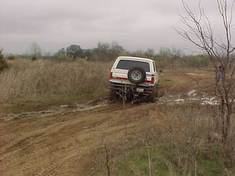 1990 Ford Bronco - A white SUV navigates a muddy off-road path, its spare tire mounted on the rear.