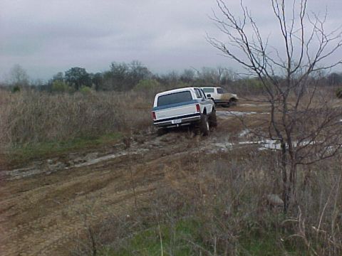 1990 Ford Bronco - Two SUVs navigate a muddy off-road course under an overcast sky.