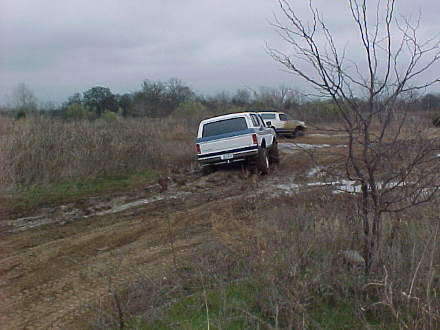 1990 Ford Bronco and another SUV driving through mud. The Bronco is white with a blue stripe