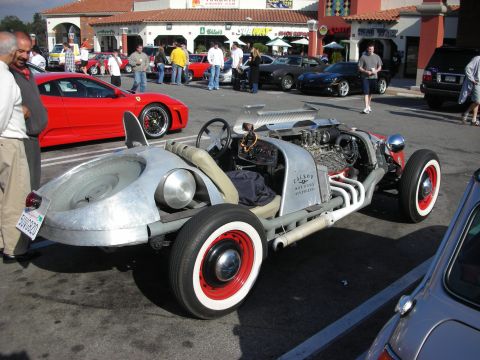 1998 Ford Crown Victoria - bare aluminum-bodied rat-rod roadster with red wire wheels and a torpedo-shaped side tank sits at a car show near a red Ferrari.