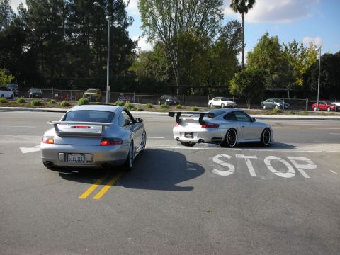 1998 Ford Crown Victoria - silver and a white Porsche 911 with rear wings parked back-to-back at an intersection marked STOP on the pavement.