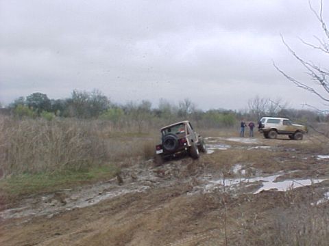 1990 Ford Bronco - Two off-road vehicles are seen navigating a challenging muddy terrain on an overcast day.