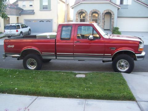 1997 Ford F-250 - A red Ford F-250 pickup truck is parked on the street in front of a suburban house, with a white SUV in the driveway.