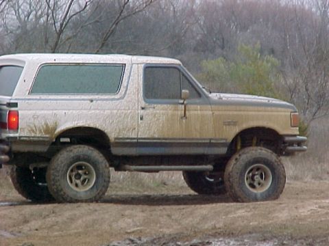 1990 Ford Bronco - Mud-covered 1990 Ford Bronco sits on a dirt path with trees in the background.