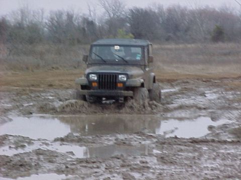 1990 Ford Bronco - A Jeep Wrangler navigates through a muddy terrain, with its front tires deep in the mire.