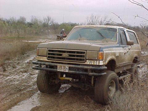 1990 Ford Bronco - This SUV is navigating a muddy terrain, while an ATV can be seen in the distance.