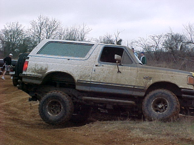 Mud-caked tan full-size Bronco with spare tire on rear, parked at an off-road event