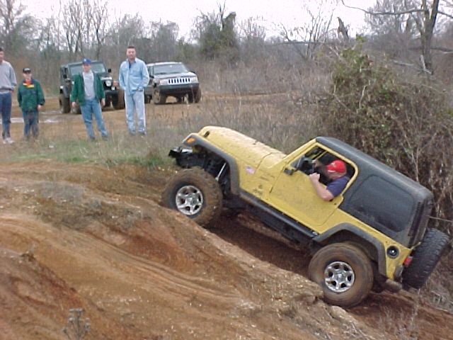 Yellow Jeep Wrangler crawling down a dirt embankment as several men watch, SUVs parked behind