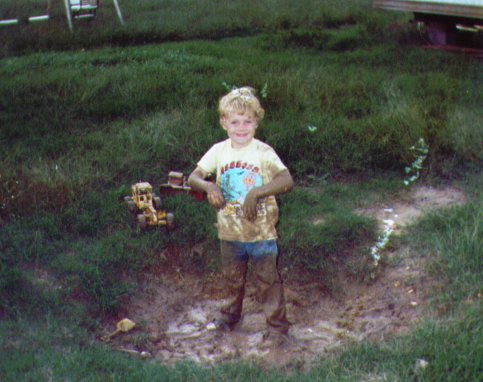 Young blond child in yellow shirt standing muddy in a grassy yard holding a toy construction truck