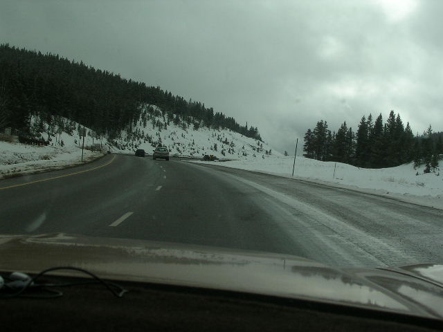 Dashboard view of a highway with snow-covered trees and two cars driving on a wet road