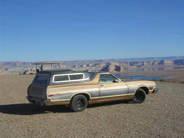 1975 Ford Ranchero with a camper shell, tan and grey, parked on a gravelly overlook with a lake and canyon in the background