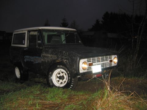 1968 Ford Bronco - A muddy 1968 Ford Bronco sits in a grassy area under the night sky, its headlights illuminating the surroundings.