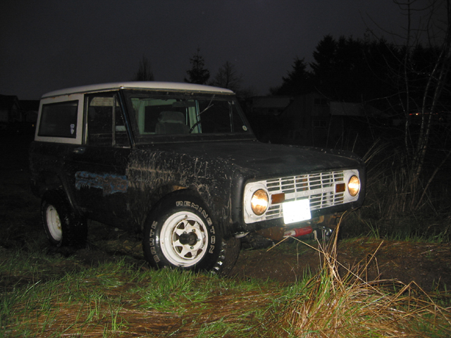Black 1968 Ford Bronco with white roof and white wheels, muddy, parked in grass at night