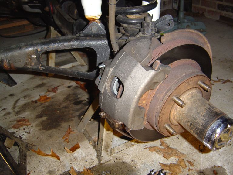 Vehicle brake caliper and rotor assembly on jack stands, with rusty metal components and fallen leaves on the ground