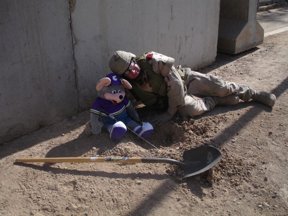 Soldier lying on the ground next to a Chuck E. Cheese plush toy, with a shovel nearby