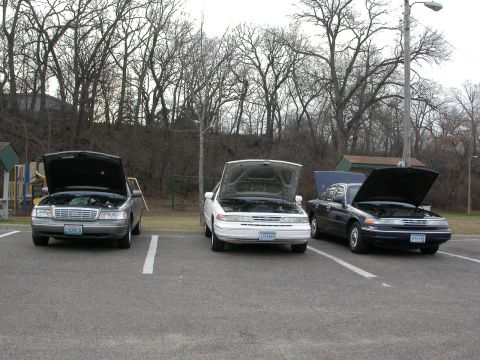 2001 Ford Crown Victoria - Three Ford Crown Victorias are parked in a lot with their hoods open, revealing their engines.