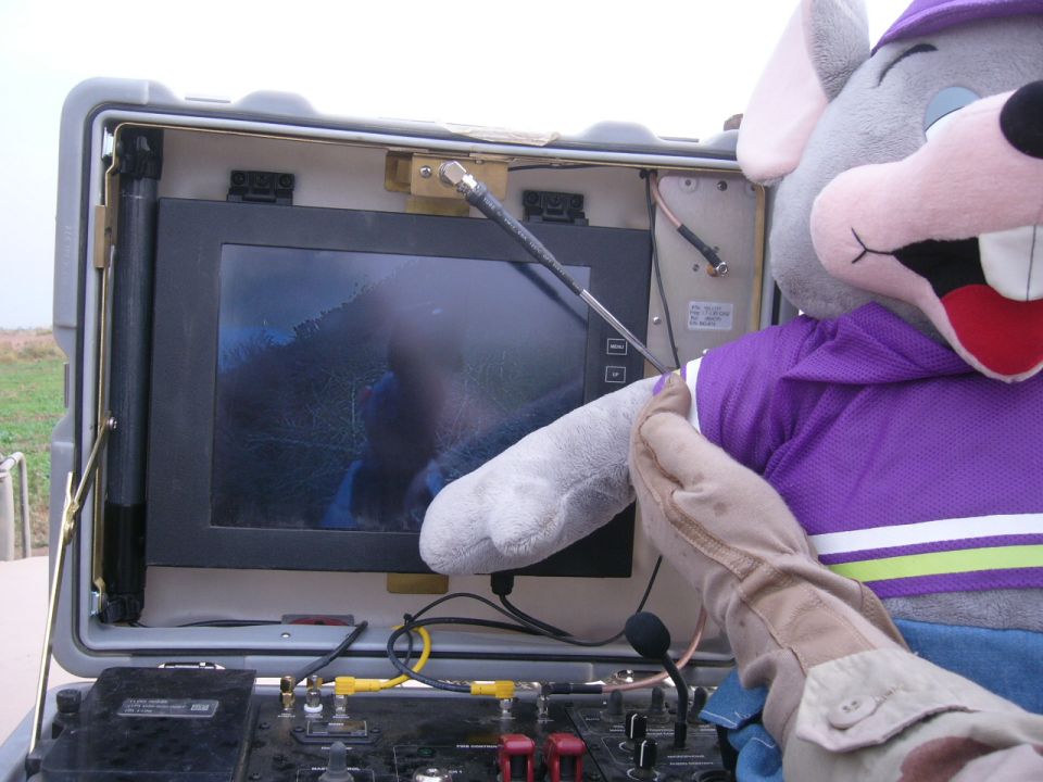 Chuck E. Cheese mascot's gloved hand touches a monitor displaying a blurry outdoor scene. Control panel visible below