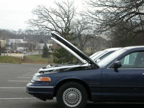 2001 Ford Crown Victoria - A dark blue 2001 Ford Crown Victoria sits in a parking lot with its hood open, alongside another vehicle.