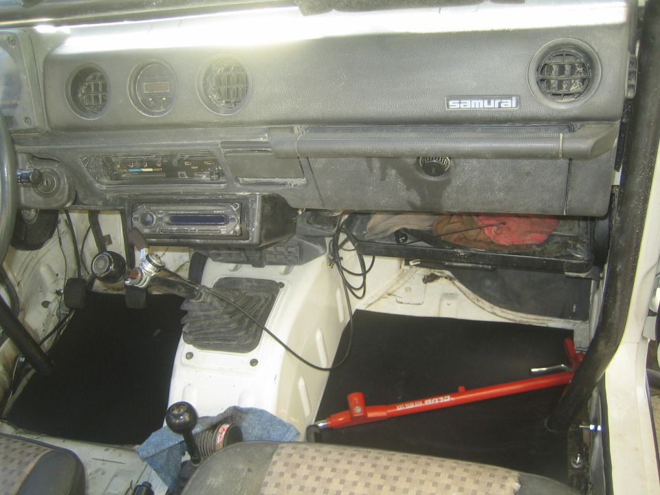Suzuki Samurai interior with dashboard, radio, gear shifter, and floor mats. A red jack is on the passenger side floor