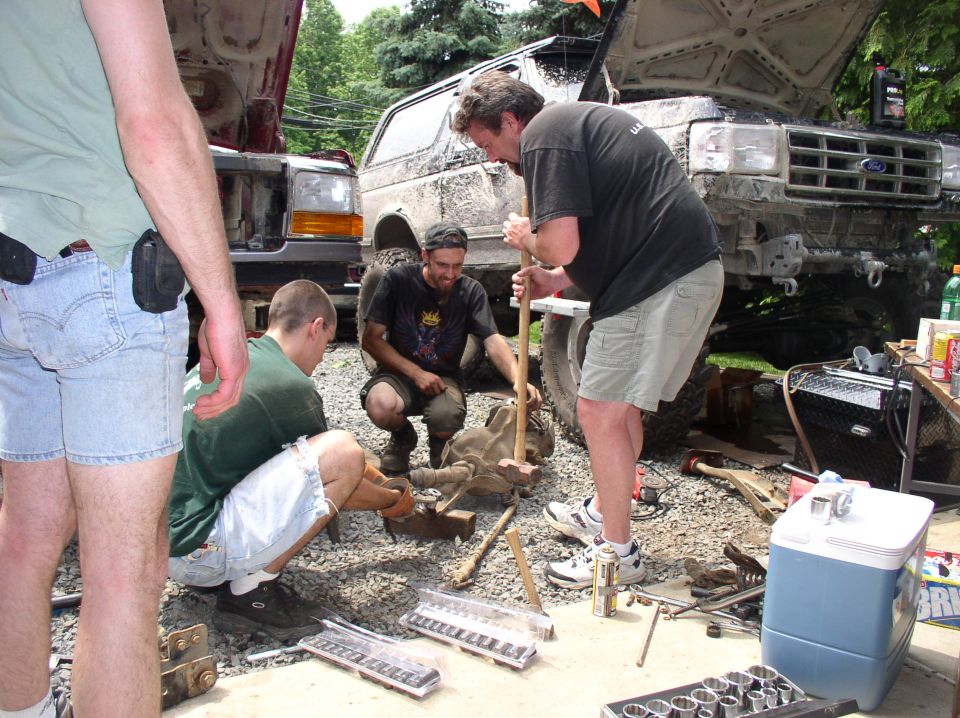 Guys working on a truck's transmission and suspension parts. Two trucks are visible