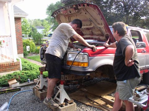 1996 Ford F-250 - Two men work on the engine of a red and silver 1996 Ford F-250 pickup truck lifted on jack stands.