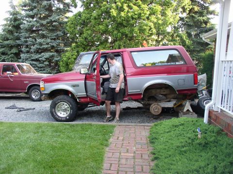 1996 Ford F-250 - A red and silver Ford Bronco is shown with its door open, lifted on jack stands in a driveway next to another maroon truck.