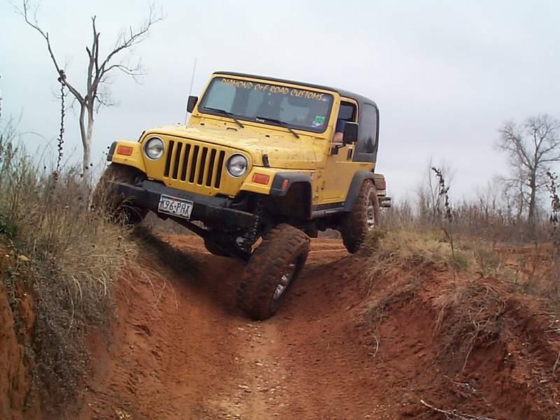 Yellow Jeep Wrangler with windshield banner straddling a deep red-clay rut on a trail