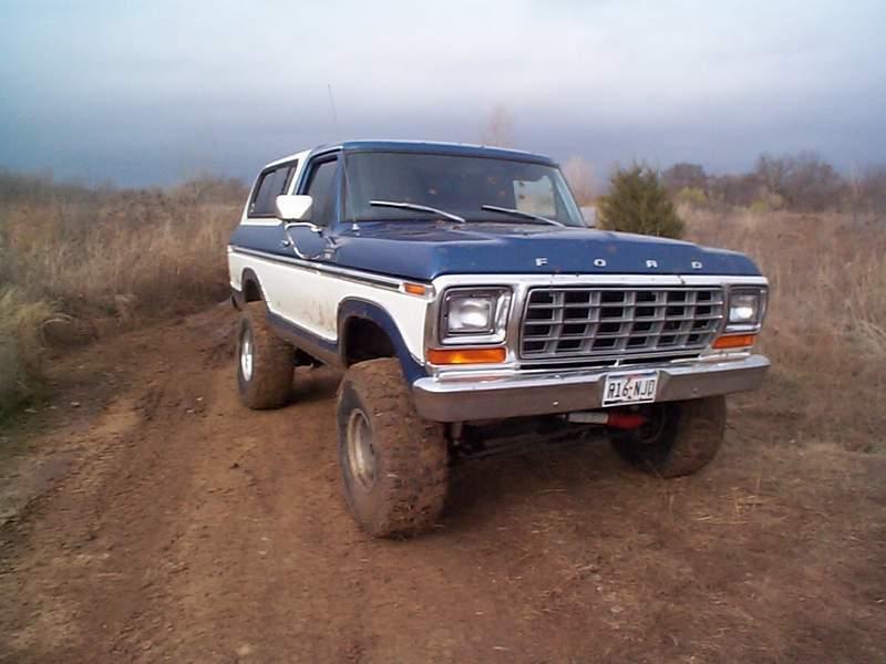Blue-and-white lifted Ford Bronco with FORD grille badge on oversized muddy tires in a field