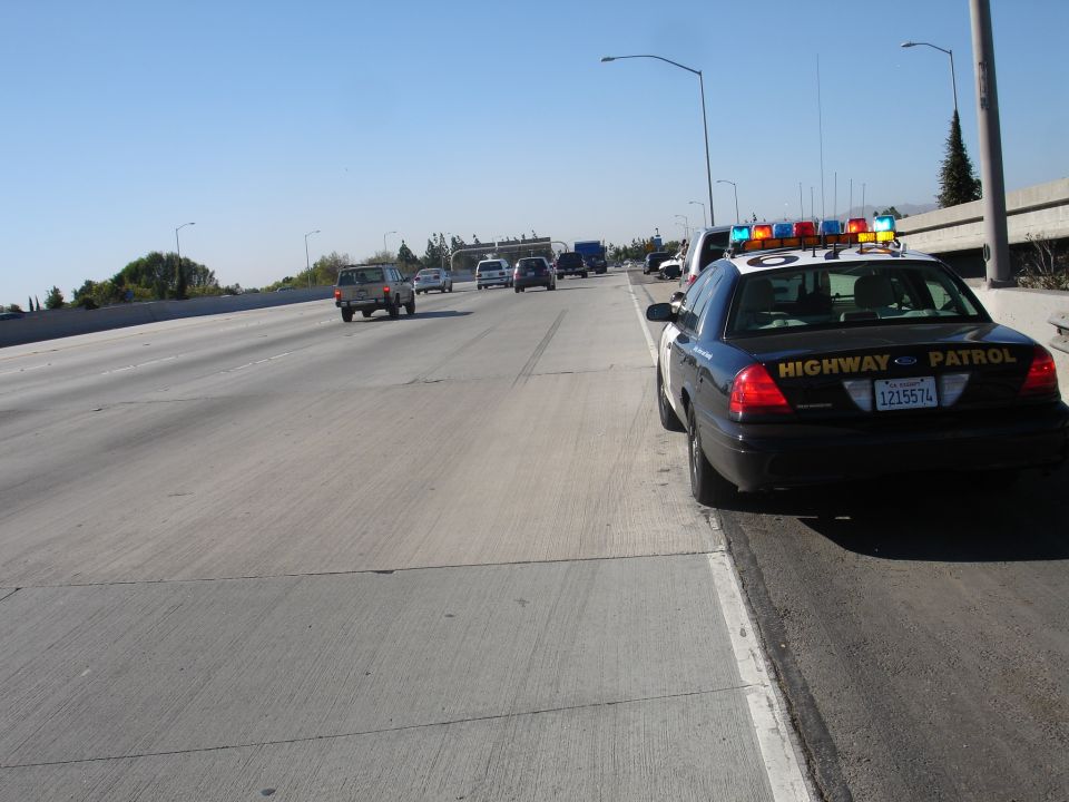 Black and white highway patrol car with flashing lights, parked on the right shoulder of a freeway