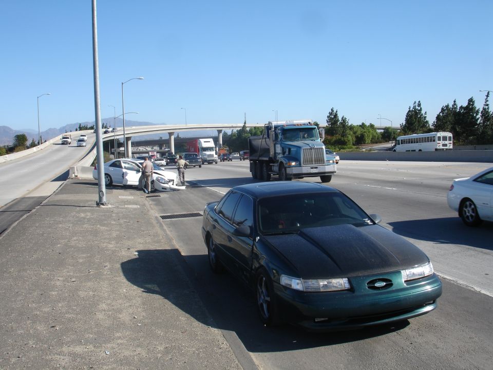 Dark green 1993 Ford Taurus with carbon fiber hood parked on highway shoulder, white car crashed ahead