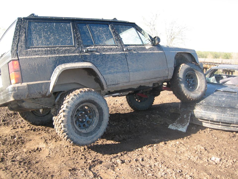 Mud-covered Jeep Cherokee XJ on its side, front tire on another car. Modified for off-road use